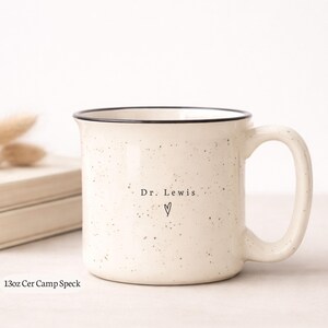 May include: Cream-colored ceramic mug with black rim and handle. The mug has a speckled design and the text "Dr. Lewis" with a heart symbol. The mug is 13oz.