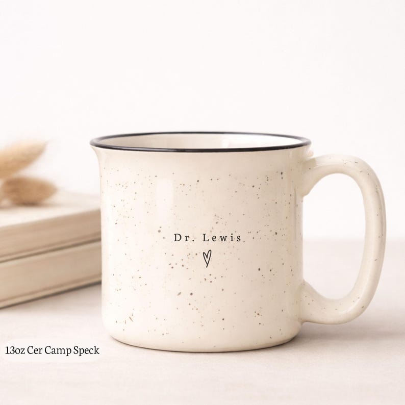 May include: Cream-colored ceramic mug with black rim and handle. The mug has a speckled design and the text "Dr. Lewis" with a heart symbol. The mug is 13oz.