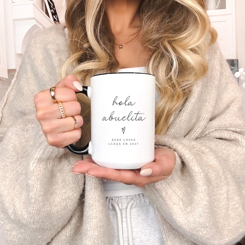 May include: White ceramic mug with a black handle and rim. The mug features the text "hola abuelita" in a handwritten style, with a heart symbol and the text "Bebe Lopez llega en 2027" below. The mug is held by a person.