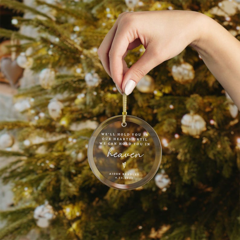 May include: A clear glass Christmas ornament with a gold string. The ornament has the text "We'll hold you in our hearts until we can hold you in heaven" with a small heart symbol. The ornament is personalized with the name "Aiden Bradley" and the date "4.15.2023".