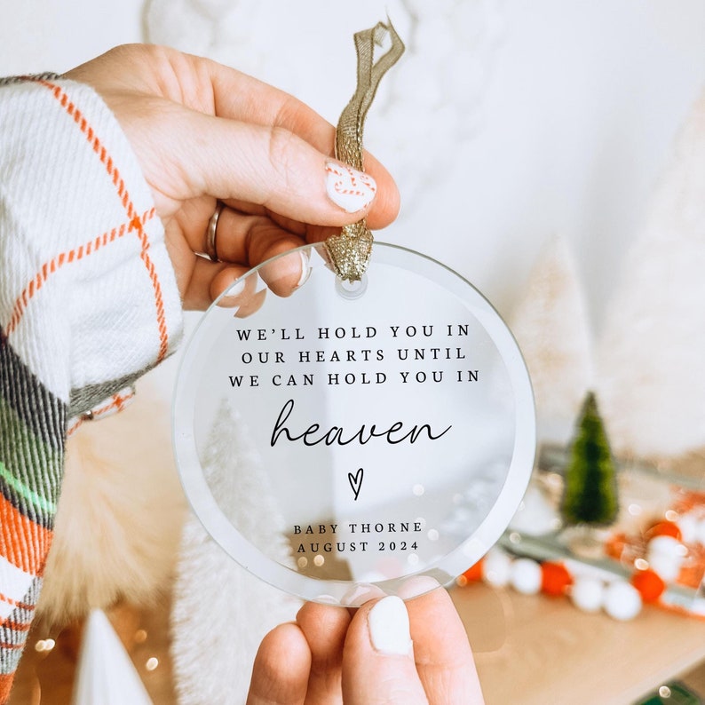 May include: A clear glass ornament with the text "We'll hold you in our hearts until we can hold you in heaven" and a heart symbol. Below the text is "Baby Thorne August 2024".