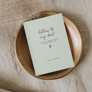 May include: A light green journal with the text "letters to my dad" and "ALL THE THINGS I WISH I COULD TELL YOU" in black script. A small heart symbol is below the text. The text "LOVE, CRISTINA" is at the bottom of the journal.