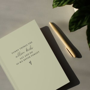 May include: A pale green journal with the text "Funny things the allen kids say and do so we never forget" and a heart symbol. A light yellow pen lies beside the journal on a grey surface.