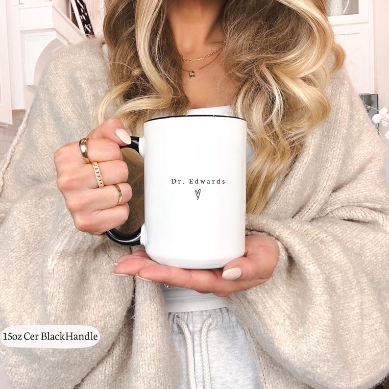 May include: White ceramic mug with a black handle and rim. The mug features the text "Dr. Edwards" and a heart symbol. The mug is held by a person wearing gold rings and a beige sweater.
