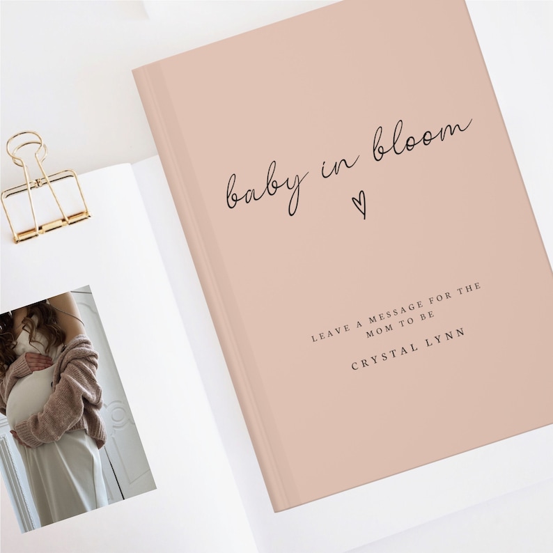 May include: A blush pink hardcover journal with the text "baby in bloom" in black cursive script and a small heart symbol. The text "Leave a message for the mom to be Crystal Lynn" is printed in black below the title.