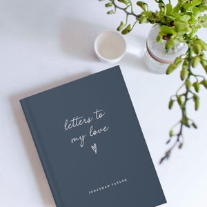 Może przedstawiać: Twardo oprawiony dziennik w ciemnoniebieskim kolorze z napisem "letters to my love" w białym kolorze i małym symbolem serca. Imię autora, "JONATHAN TAYLOR", jest nadrukowane na dole okładki białym kolorem.