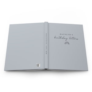 May include: A light gray journal with the text "KATELYN'S birthday letters" and a rainbow graphic. The spine reads "DEAR KATELYN | LOVE, MOM". A small barcode is visible on the back cover. The journal is open, displaying the inside pages.