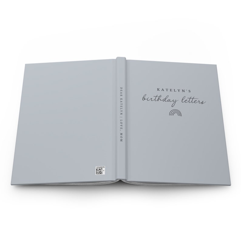 May include: A light gray journal with the text "KATELYN'S birthday letters" and a rainbow graphic. The spine reads "DEAR KATELYN | LOVE, MOM". A small barcode is visible on the back cover. The journal is open, displaying the inside pages.