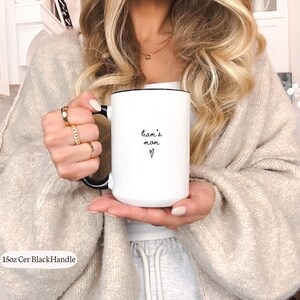 May include: A white ceramic mug with a black handle and rim, featuring the black text 'liam's mom' and a small heart symbol printed on the front. The mug is labeled as 15oz, indicating its capacity, and has a smooth, glossy finish.