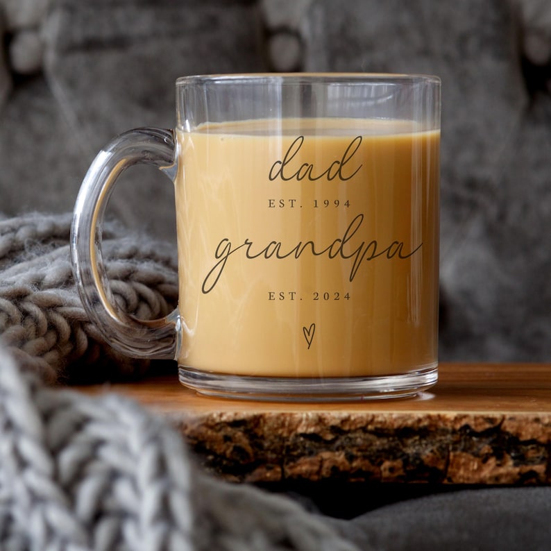 May include: A clear glass mug filled with a light brown liquid, featuring the words "dad est. 1994" and "grandpa est. 2024" in elegant script. A small heart symbol is below the text. The mug has a clear handle.