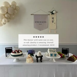 May include: A white table with a white tablecloth, decorated with a banner that reads "EMERSON" in Korean and English, and a banner that reads "백일" in Korean. There are several white cake stands with desserts on them, including a cake with a "Happy Birthday" sign, a stack of white cubes with blue writing, and a bouquet of purple flowers. The table is decorated with a cluster of white balloons.