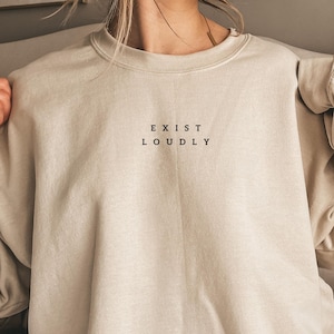Peut inclure: Un sweat-shirt à col rond beige avec le texte noir "EXIST LOUDLY" imprimé dans une police sans empattement sur le centre de la poitrine. Le vêtement présente des manches longues, un col côtelé et un design simple de couleur unie.
