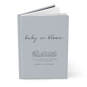 May include: A light gray hardcover book with the title "baby in bloom" in black cursive script. The book has a black and white illustration of flowers and the text "Leave a message for the future parents Anne & Michael".