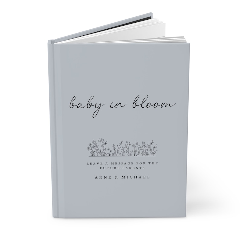May include: A light gray hardcover book with the title "baby in bloom" in black cursive script. The book has a black and white illustration of flowers and the text "Leave a message for the future parents Anne & Michael".
