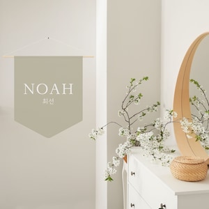 May include: A light beige fabric banner with the word "NOAH" and the Korean word "최선" written in white. The banner is hanging from a wooden dowel rod and is displayed on a white wall.
