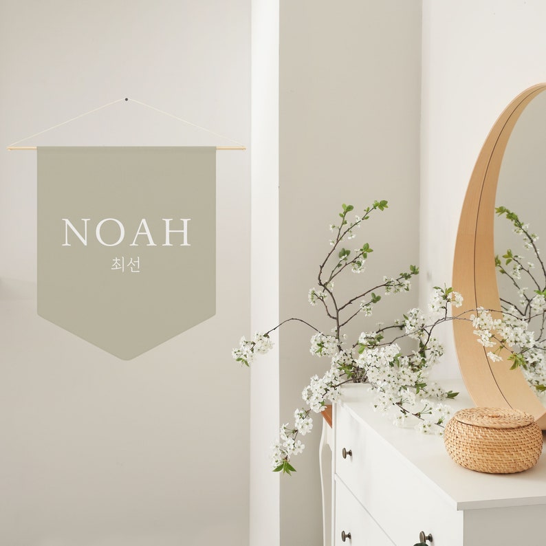 May include: A light beige fabric banner with the word "NOAH" and the Korean word "최선" written in white. The banner is hanging from a wooden dowel rod and is displayed on a white wall.