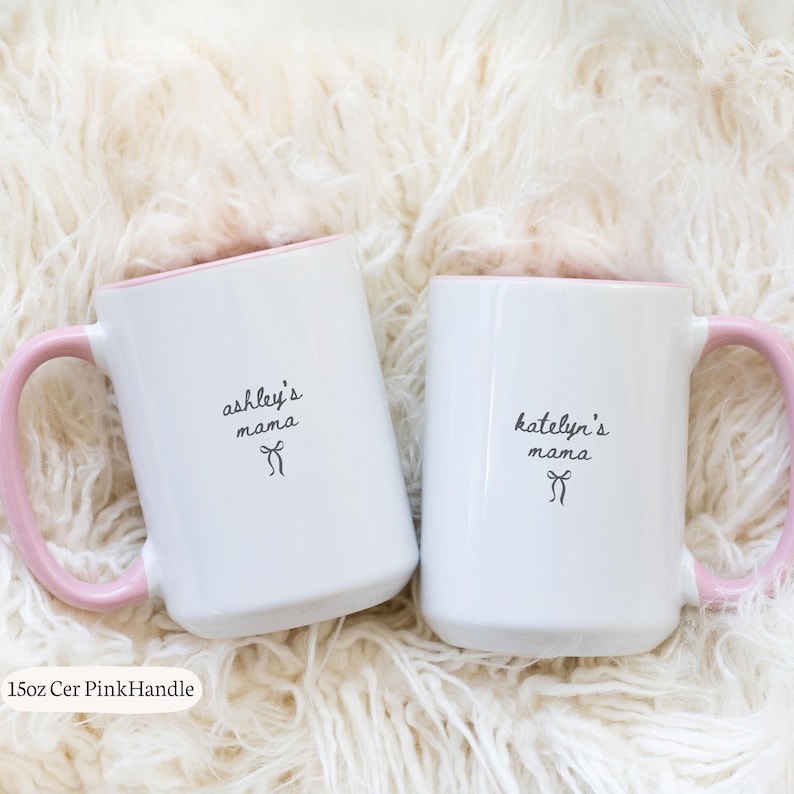 May include: Two white ceramic mugs with pink handles, each featuring a 15 oz capacity. One mug is printed with the text ashley's mama and a small bow icon, while the other is printed with katelyn's mama and a small bow icon in black ink.