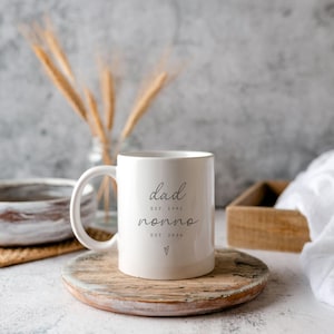 May include: A white ceramic mug with the words "dad nonno" in a handwritten font. The mug includes the dates "EST. 1991" and "EST. 2024" with a small heart symbol. The mug sits on a wooden coaster.