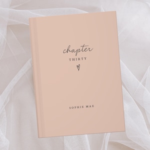 Op de afbeelding: Een perzikroze hardcover-dagboek met de woorden "chapter THIRTY" en een hartsymbool in zwart schrift. De naam "SOPHIE MAE" staat eronder gedrukt. Het dagboek ligt op een witte, doorschijnende stoffen achtergrond.
