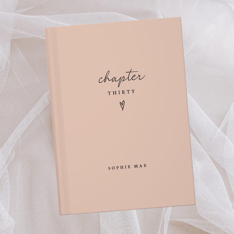 Op de afbeelding: Een perzikroze hardcover-dagboek met de woorden "chapter THIRTY" en een hartsymbool in zwart schrift. De naam "SOPHIE MAE" staat eronder gedrukt. Het dagboek ligt op een witte, doorschijnende stoffen achtergrond.
