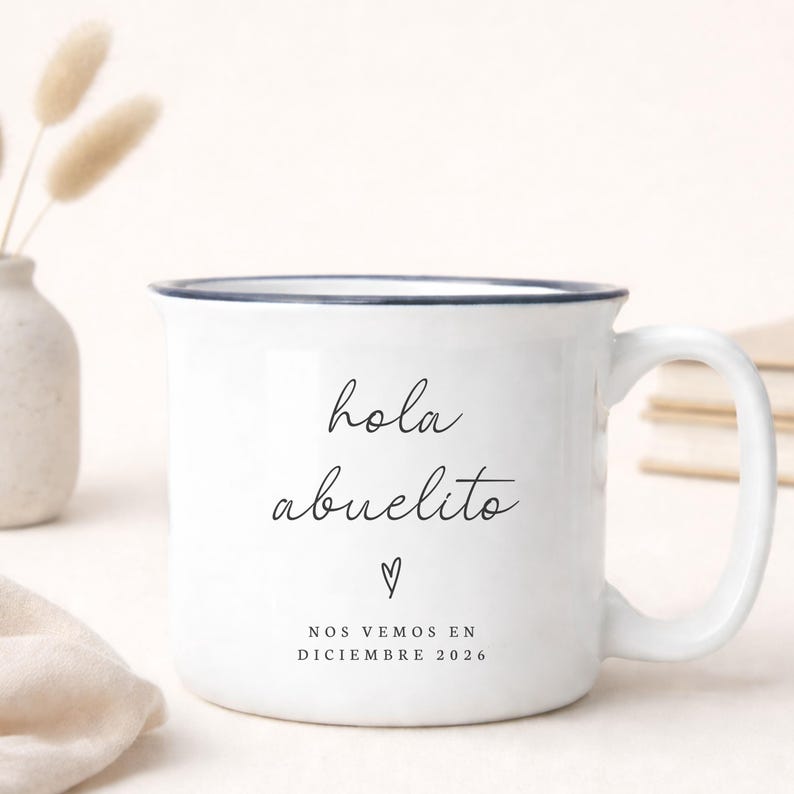 May include: White ceramic mug with a blue rim and handle. The mug features the text "hola abuelito" in black script, a heart symbol, and the text "NOS VEMOS EN DICIEMBRE 2026". A small vase with dried flowers is in the background.