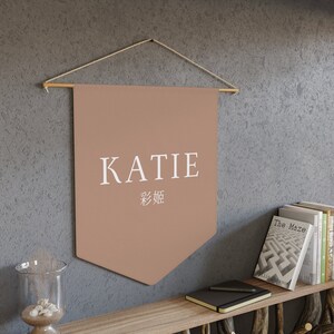 May include: A brown pennant banner with the name "KATIE" in white text and the Chinese characters for "Katie" below. The banner is hanging from a wooden dowel rod with twine.
