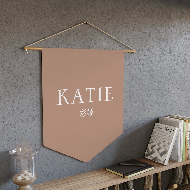 May include: A brown pennant banner with the name "KATIE" in white text and the Chinese characters for "Katie" below. The banner is hanging from a wooden dowel rod with twine.
