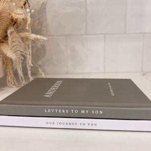 May include: Two hardcover books stacked on top of each other. The top book is gray with the name "ANDERSON" in white lettering. The title "LETTERS TO MY SON" is below. The bottom book is white with the title "OUR JOURNEY TO YOU".