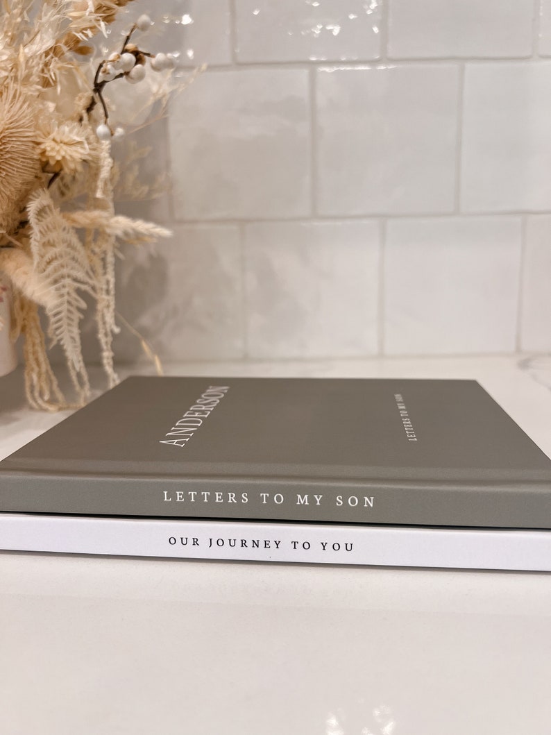 May include: Two hardcover books stacked on top of each other. The top book is gray with the name "ANDERSON" in white lettering. The title "LETTERS TO MY SON" is below. The bottom book is white with the title "OUR JOURNEY TO YOU".