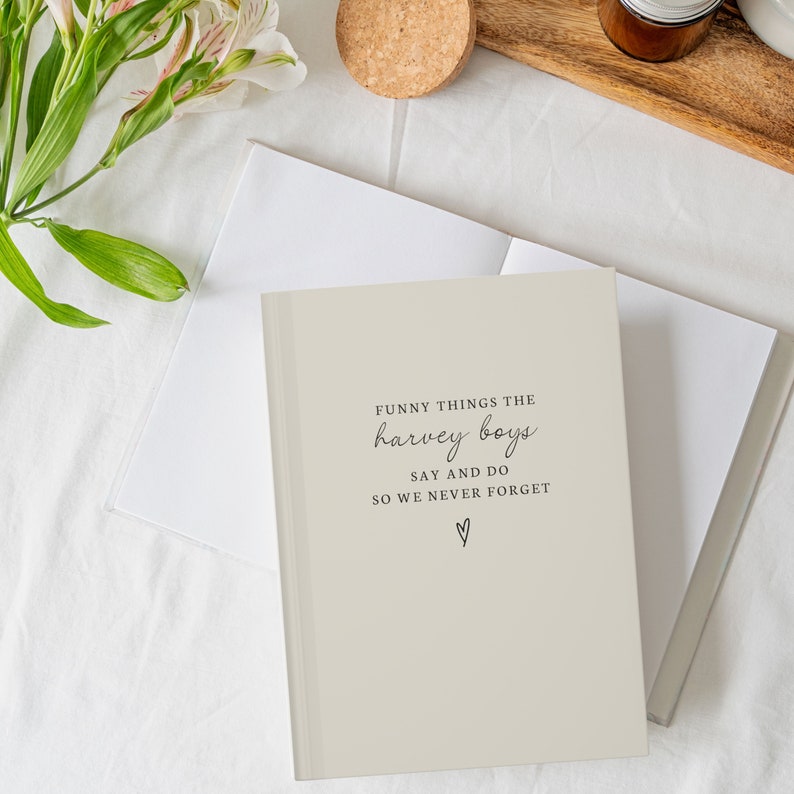 May include: A beige journal with the text "FUNNY THINGS THE harvey boys SAY AND DO SO WE NEVER FORGET" in black script. The journal is open to reveal blank white pages. A small heart symbol is below the text. The journal is on a white surface.