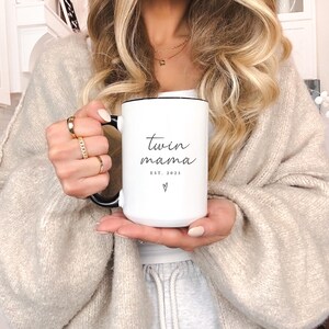 May include: White ceramic mug with a black handle and rim. The mug features the text "twin mama" in a script font, with "EST. 2025" and a heart symbol below. The mug is held by a person.