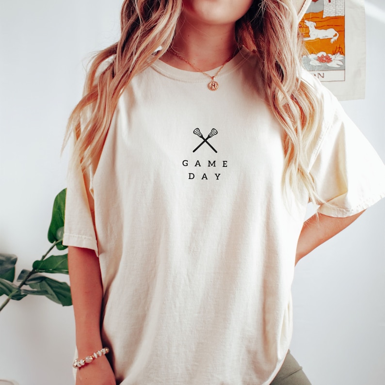 May include: A cream colored t-shirt with a black graphic of two lacrosse sticks crossed with the text "GAME DAY" underneath.