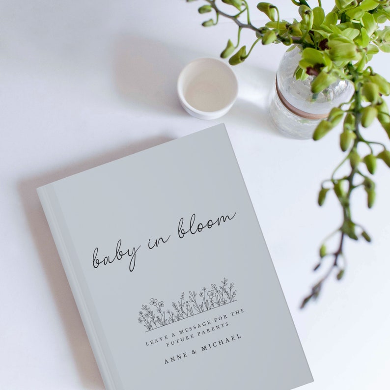 May include: A gray book with the text "baby in bloom" and a floral design. The text "Leave a message for the future parents" and "Anne & Michael" is printed below the floral design.