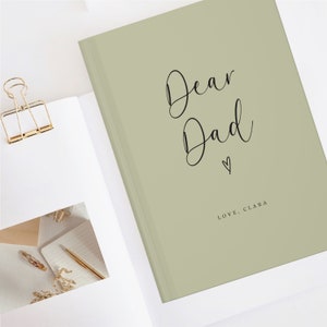May include: A sage green hardcover book with the black cursive text Dear Dad and a small heart icon printed on the front cover. Below the heart, the text reads LOVE, CLARA in a simple sans-serif font. The book is positioned on a white surface.