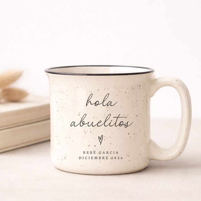 May include: Cream-colored ceramic mug with black speckled design and a black rim. The mug features the text "hola abuelitos" in a handwritten font, a small heart, and the text "BEB&Eacute; GARCIA DICIEMBRE 2026".