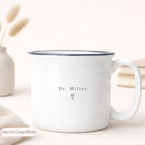 May include: White ceramic camp mug with a navy blue rim and handle. The mug features the text "Dr. Miller" and a heart symbol in black. The mug is labeled "13oz Cer Camp White".