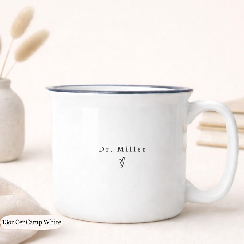 May include: White ceramic camp mug with a navy blue rim and handle. The mug features the text "Dr. Miller" and a heart symbol in black. The mug is labeled "13oz Cer Camp White".