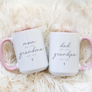 May include: Two white ceramic mugs with pink handles and rims. One mug reads "mom grandma EST. 1993 EST. 2024 ♡" and the other reads "dad grandpa EST. 1993 EST. 2024 ♡". The mugs are on a fluffy white surface.