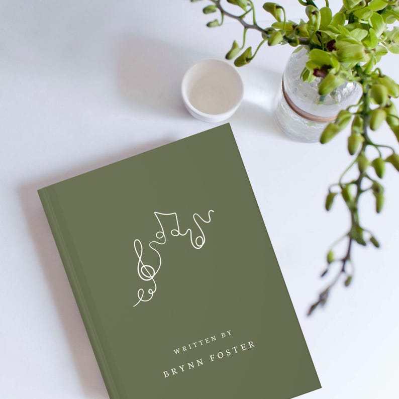 May include: An olive green book with a white line drawing of musical notes and a treble clef. The text "WRITTEN BY BRYNN FOSTER" is printed in white at the bottom of the book. A small white cup and a glass vase with green plants are in the background.