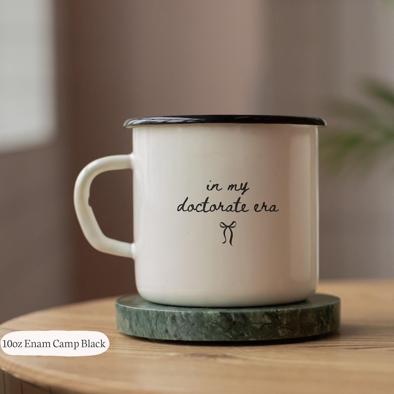 May include: A white enamel camp mug with a black rim and handle, resting on a green marble coaster. The mug features the text "in my doctorate era" in black script, with a small bow graphic below. The mug holds 10oz.