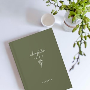 Op de afbeelding: Een olijfgroen hardcover boek met het woord "chapter" in cursief schrift. Daaronder staan het woord "THIRTY" en een bloemenillustratie in het wit gedrukt. De naam "KATHRYN" staat onderaan gedrukt. Een klein wit kopje en een glazen vaas met groen staan op de achtergrond.