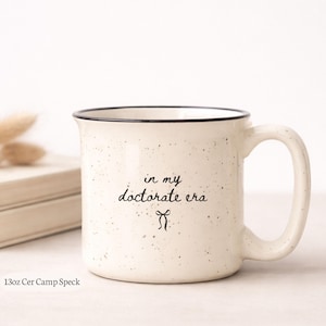 May include: Cream-colored ceramic mug with black speckled design and a black rim. The mug features the text "in my doctorate era" in a handwritten style with a small bow graphic. The mug is approximately 13 ounces.