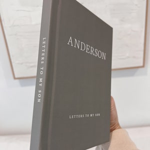May include: A gray hardcover book titled "ANDERSON" with "LETTERS TO MY SON" printed on the spine and cover. The book is held at an angle, showcasing the title in a clean, sans-serif font.