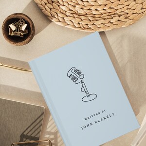 May include: A light blue book with a black line drawing of a microphone and the text "WRITTEN BY JOHN BLAKELY". A small wooden bowl with gold paper clips is in the upper left corner. A gold pen and a woven basket are also visible.