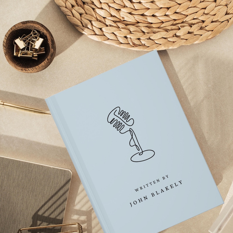 May include: A light blue book with a black line drawing of a microphone and the text "WRITTEN BY JOHN BLAKELY". A small wooden bowl with gold paper clips is in the upper left corner. A gold pen and a woven basket are also visible.