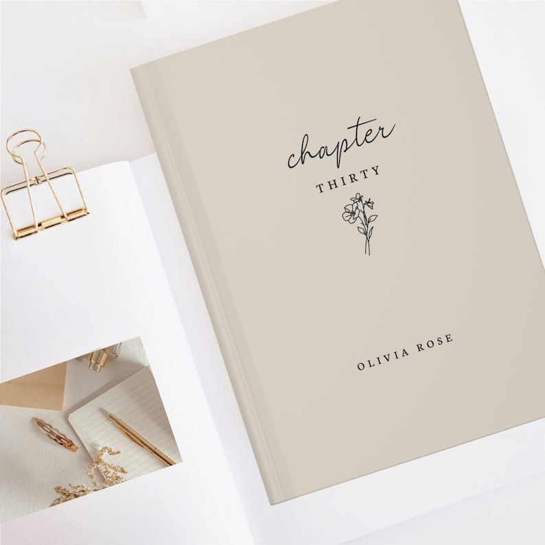 Op de afbeelding: Een beige hardcover notitieboek met de tekst "chapter thirty" en een bloemenillustratie. De naam "Olivia Rose" staat onderaan. Een gouden paperclip en andere briefpapierartikelen zijn zichtbaar.