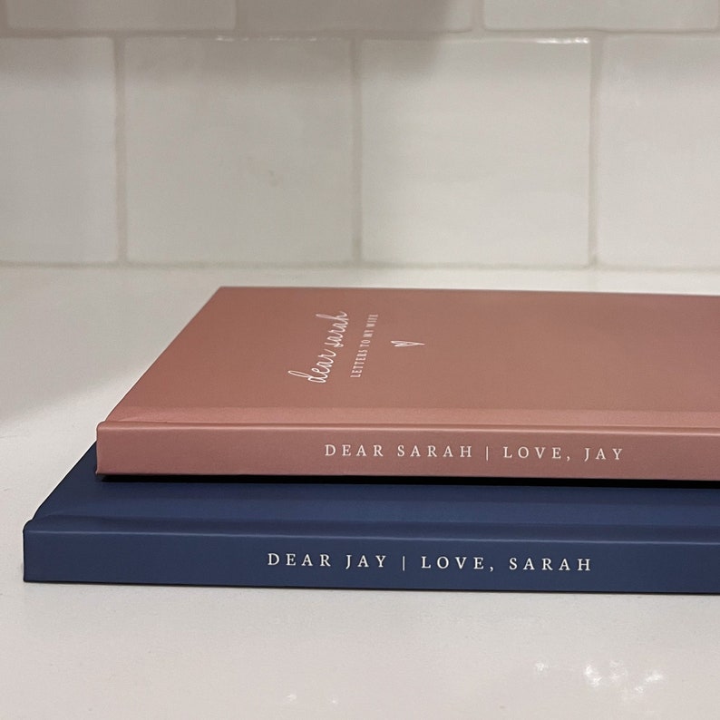 May include: Two hardcover journals, one rose-gold and one navy blue, stacked on top of each other. The rose-gold journal has the text "dear sarah" and "LETTERS TO MY WIFE". The navy blue journal has the text "DEAR JAY | LOVE, SARAH".