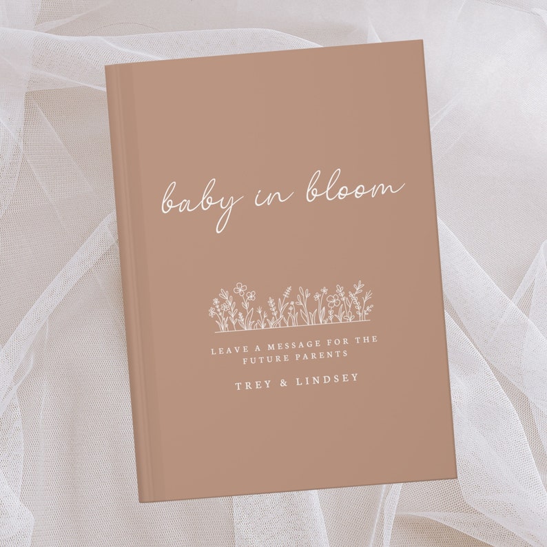 May include: A light brown book with the title "baby in bloom" in white script. The book has a white floral design and the text "Leave a message for the future parents Trey & Lindsey".