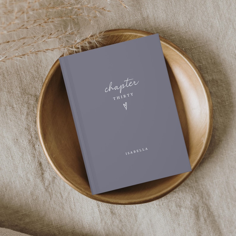 Op de afbeelding: Een grijs hardcover boek met de woorden "chapter THIRTY" en de naam "ISABELLA" in witte letters. Een klein hartsymbool staat onder het woord "THIRTY". Het boek ligt in een houten kom.