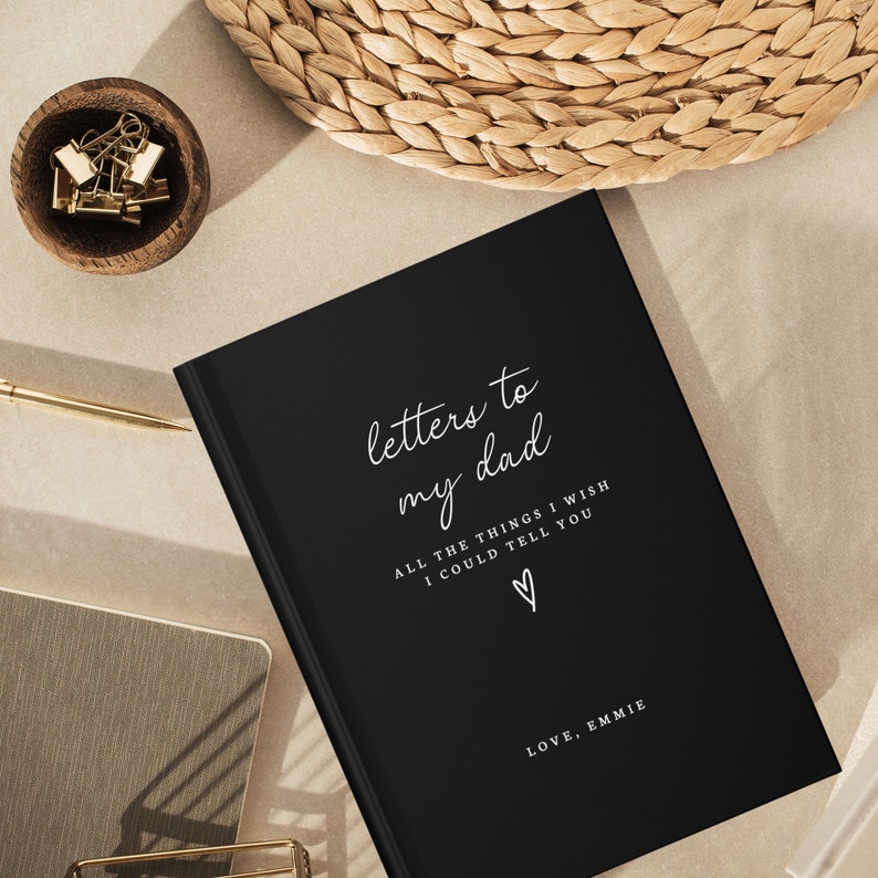 May include: A black journal with the text "letters to my dad" in white script. The text "ALL THE THINGS I WISH I COULD TELL YOU" is also in white script. A small heart symbol is below the text. The text "LOVE, EMMIE" is at the bottom of the journal.